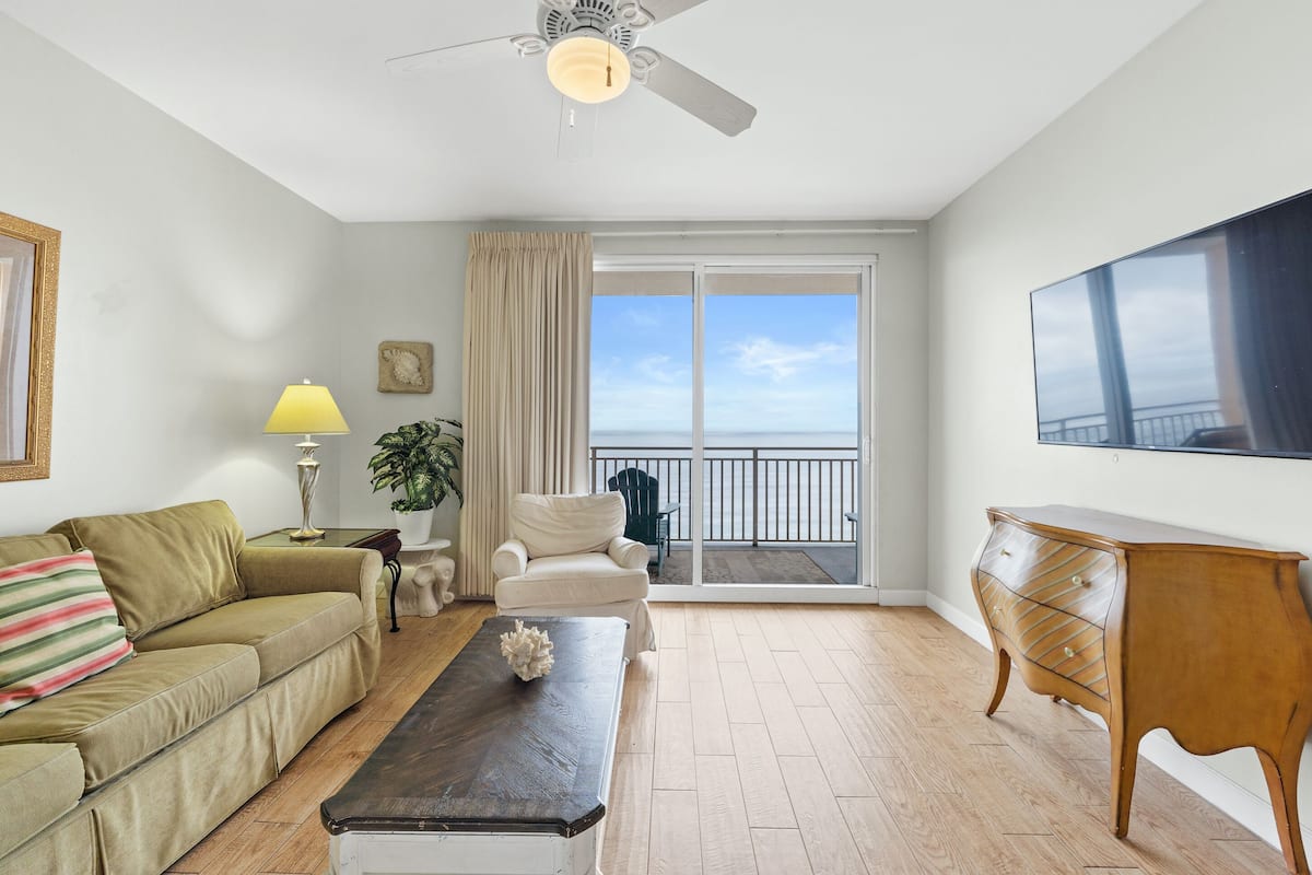 A living room with a sofa, a coffee table, a chair, a TV, a ceiling fan, and a balcony with a view.
