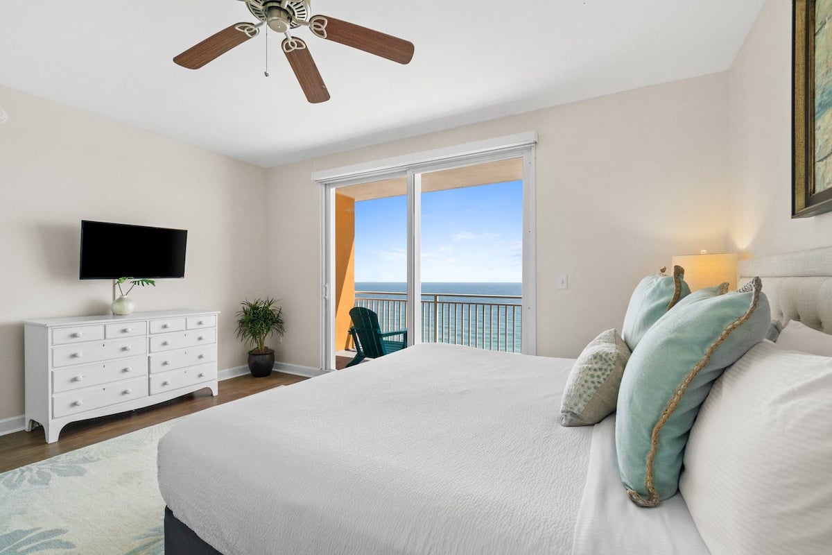 A bedroom with a large bed, a ceiling fan, a TV on a dresser, a potted plant, and a balcony with a view of the ocean.