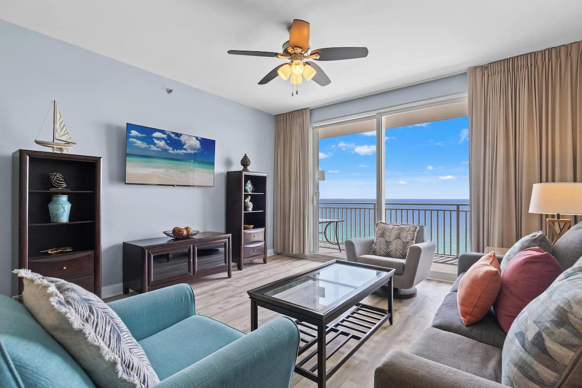 A living room with a large window, a ceiling fan, and a view of the ocean.