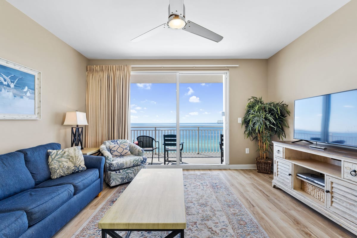 A living room with a sofa, a coffee table, a TV, and a balcony with a view of the ocean.