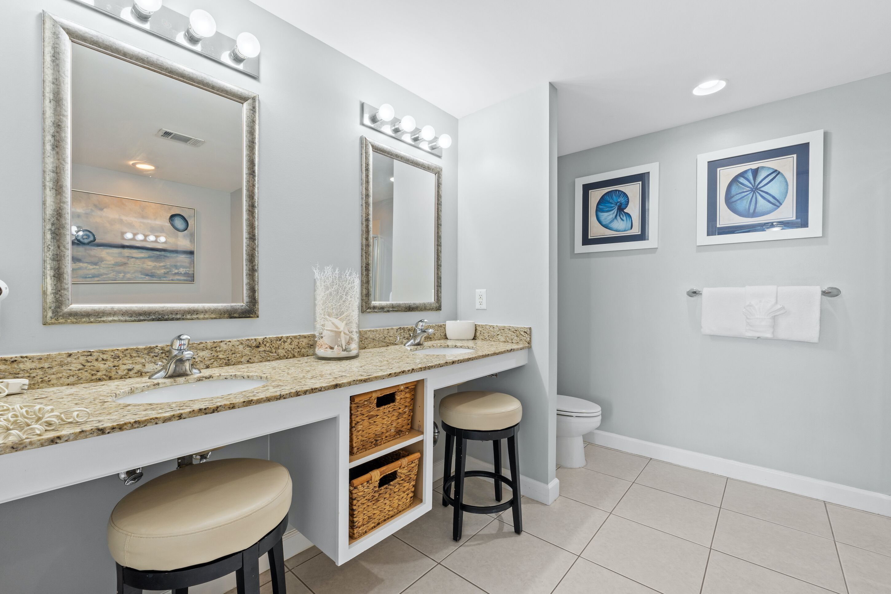 A bathroom with two sinks, a vanity with a mirror, and a toilet. There are wall-mounted lights above the mirrors and a towel rack on the wall.