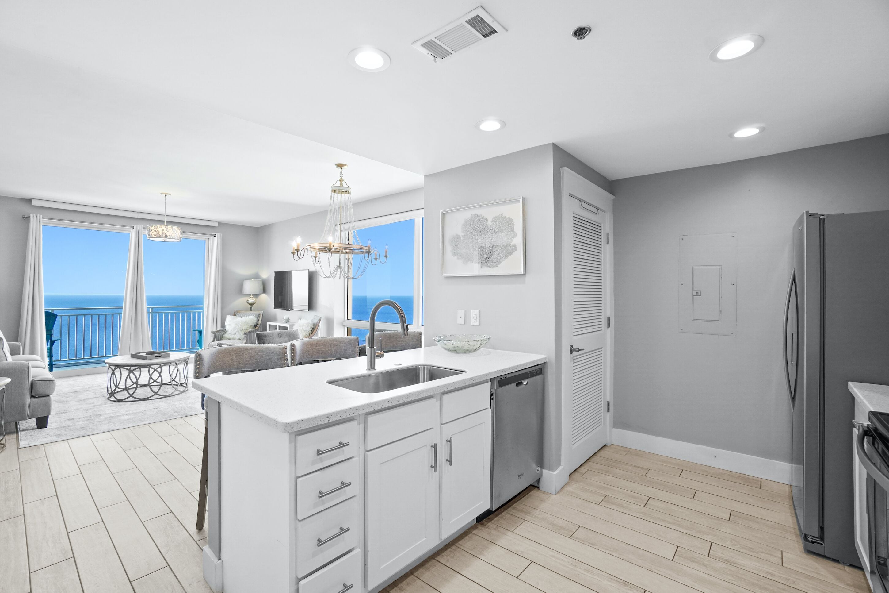 A modern kitchen with a large island, built-in dishwasher, and a refrigerator. There is a dining area with a chandelier and a view of the ocean.