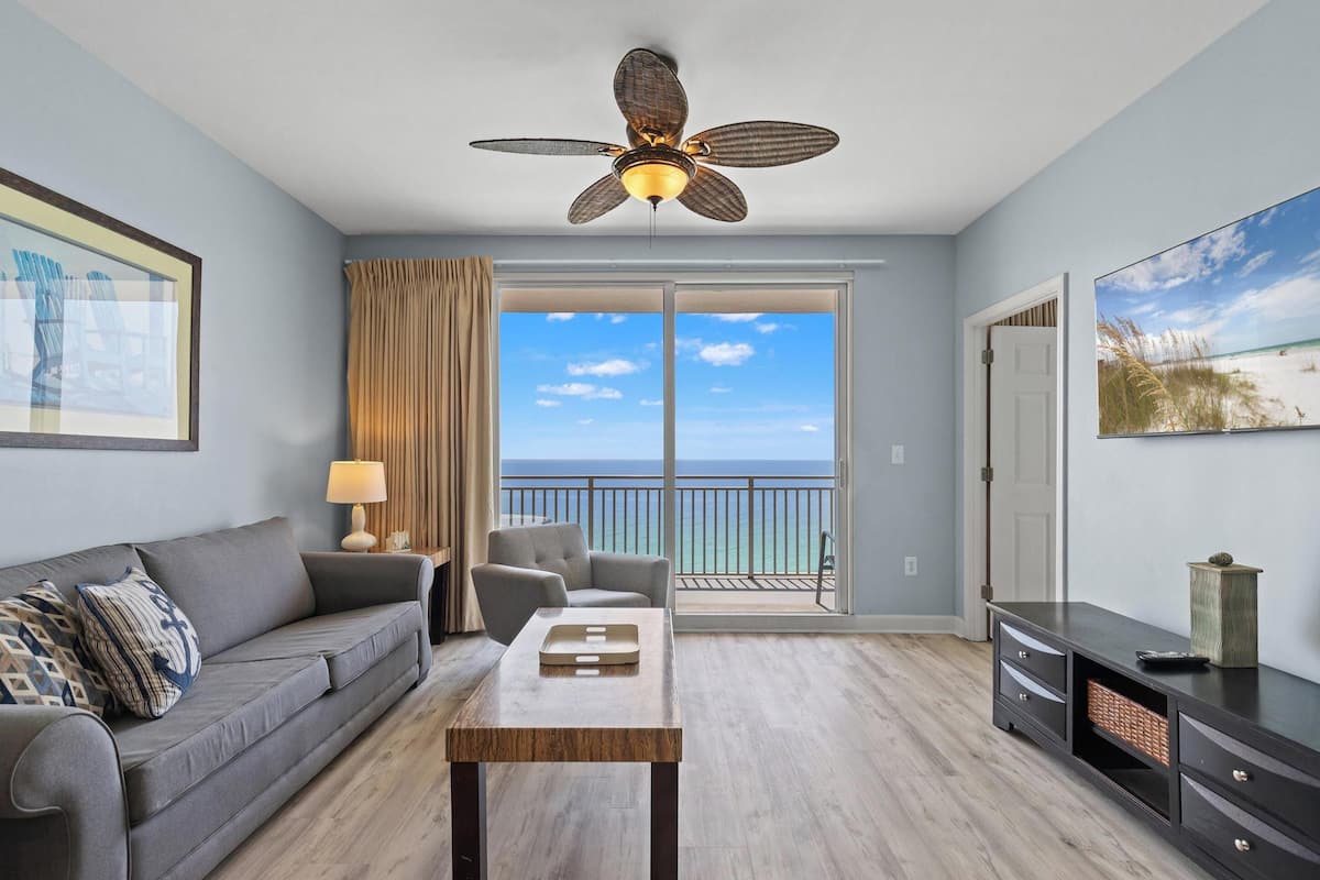 A room with a ceiling fan, a sofa, a table, a TV, and a balcony with a view of the ocean.