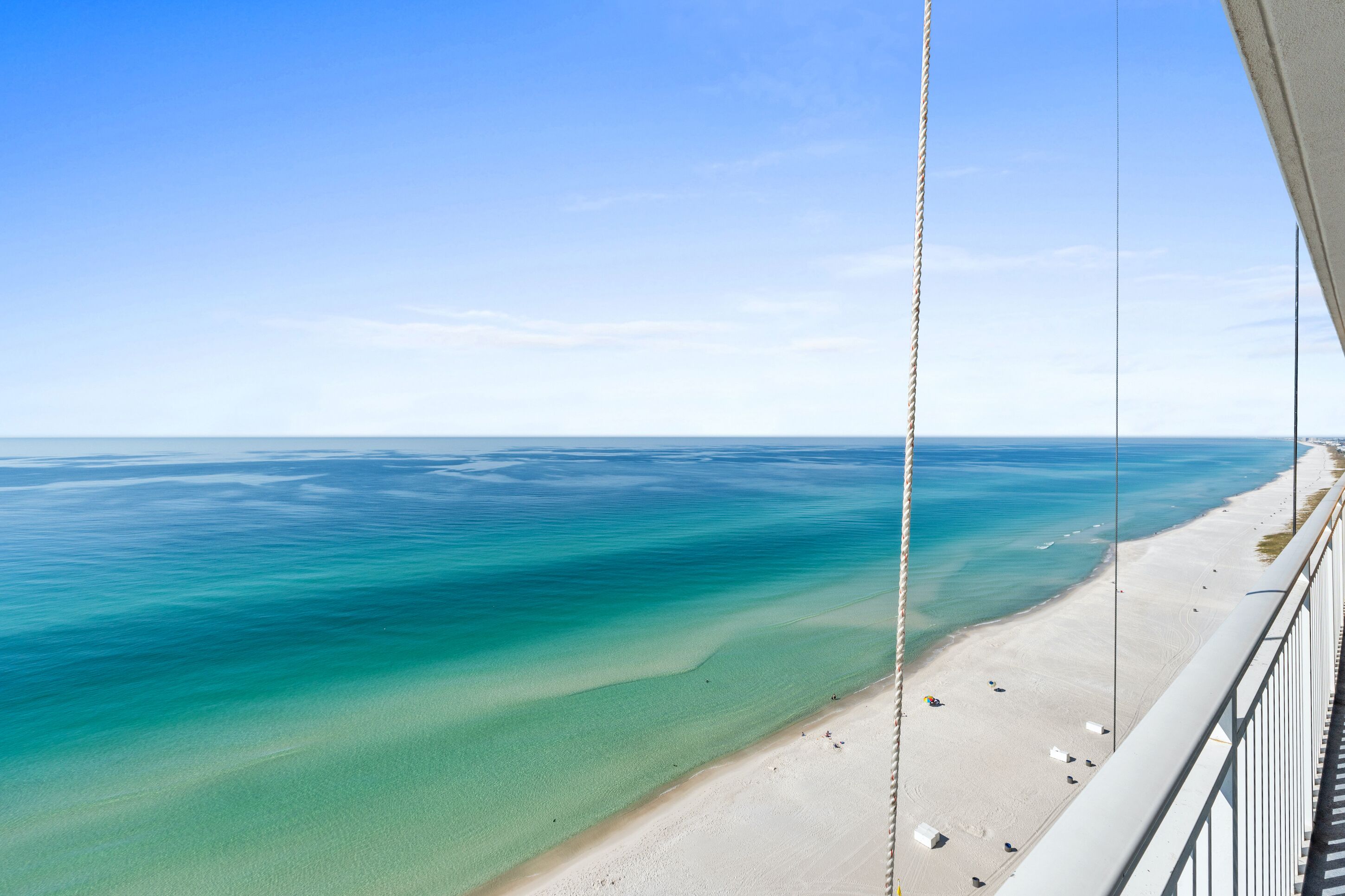 A beach view from a balcony with clear water, a sandy shore, and a rope railing.