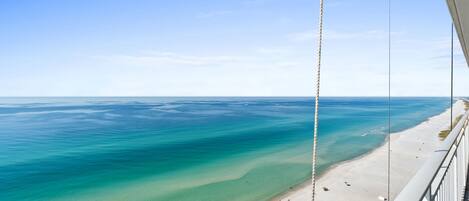 A beach view from a balcony with clear water, a sandy shore, and a rope railing.