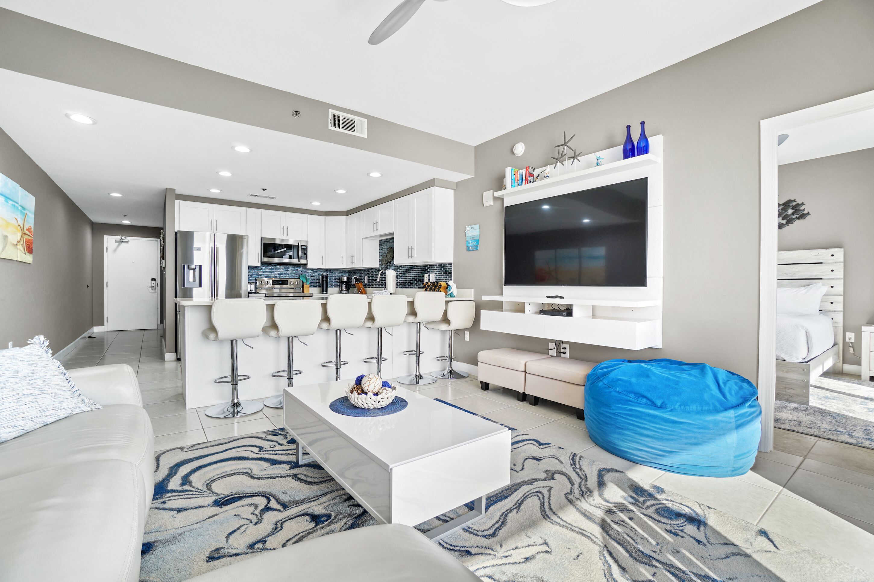A modern living room with a white sofa, a coffee table, and a blue bean bag. There is a kitchen area in the background with white cabinets and a large TV mounted on the wall.