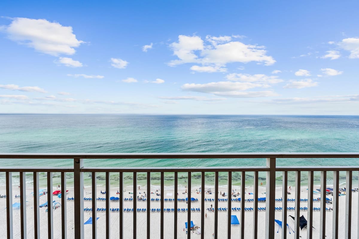 A beach view from a balcony with a railing, clear blue sky, and turquoise sea.