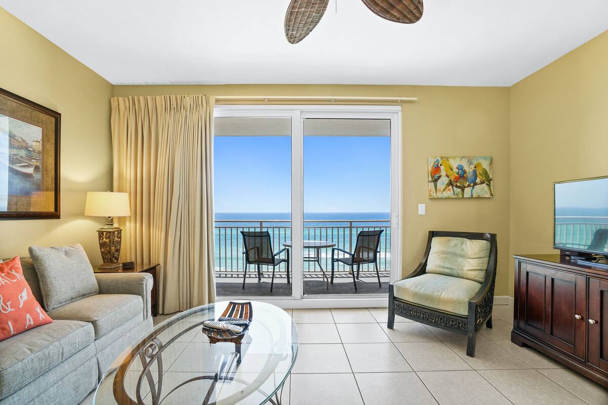 A living room with a sofa, armchair, glass coffee table, and a TV. There is a balcony with chairs and a view of the ocean.
