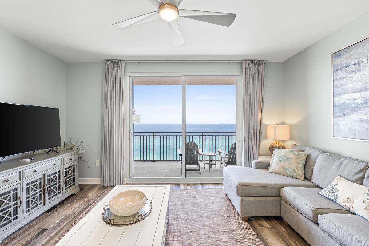 A living room with a flat-screen TV, a grey sofa, and a balcony with a view of the ocean.