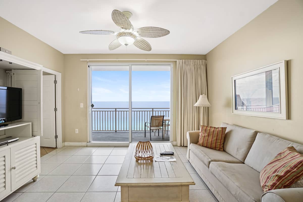 A spacious living room with a large sliding glass door leading to a balcony, a ceiling fan, a flat-screen TV, a sofa, and a coffee table.