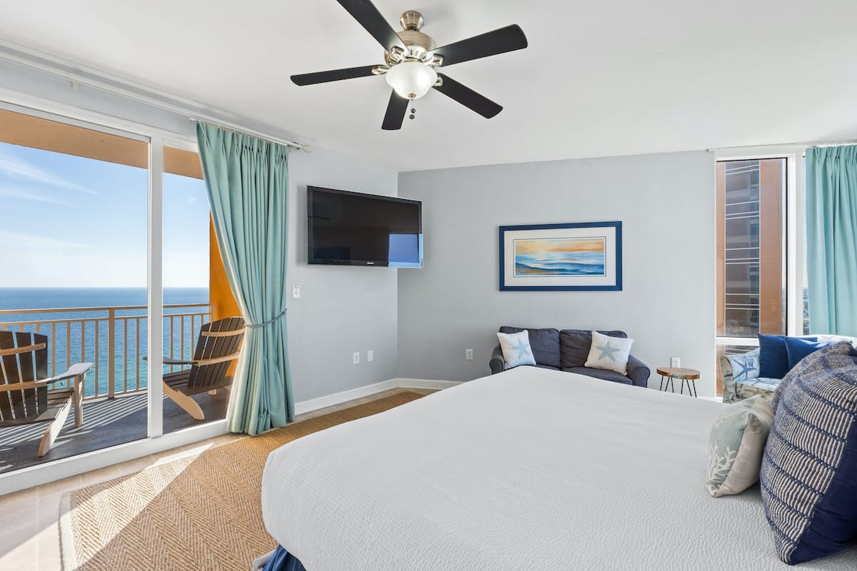 A hotel room with a large bed, a ceiling fan, a TV, a painting, and a balcony with a view of the ocean.