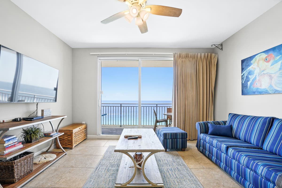 A living room with a sofa, a coffee table, a TV, and a balcony with a view of the ocean.