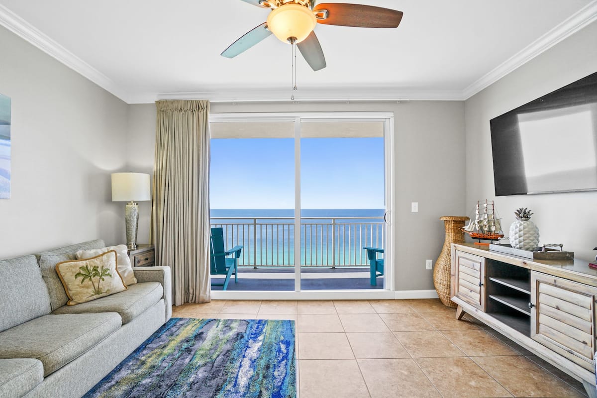 A room with a grey sofa, a blue patterned rug, a ceiling fan, a TV, a decorative ship model, and a view of the ocean through a sliding door.