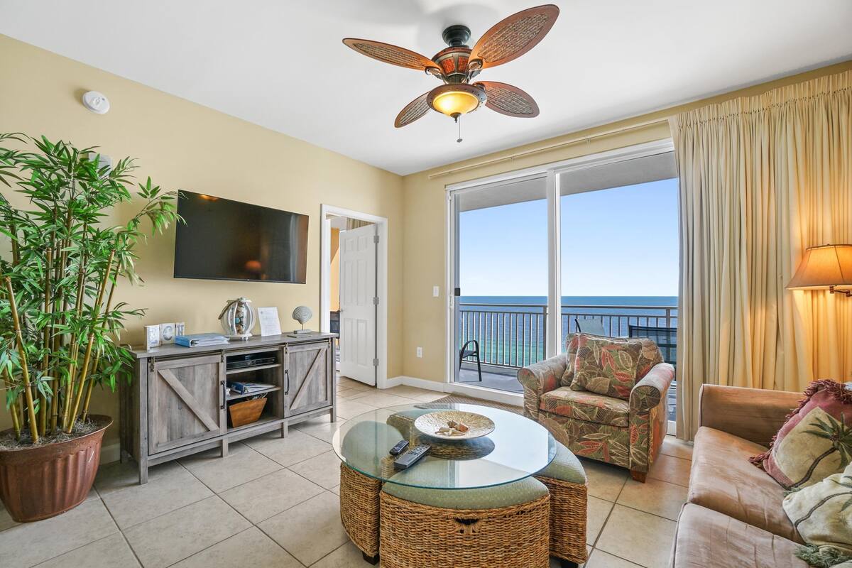 A living room with a glass coffee table, wicker furniture, a television, and a large window with a view of the ocean.