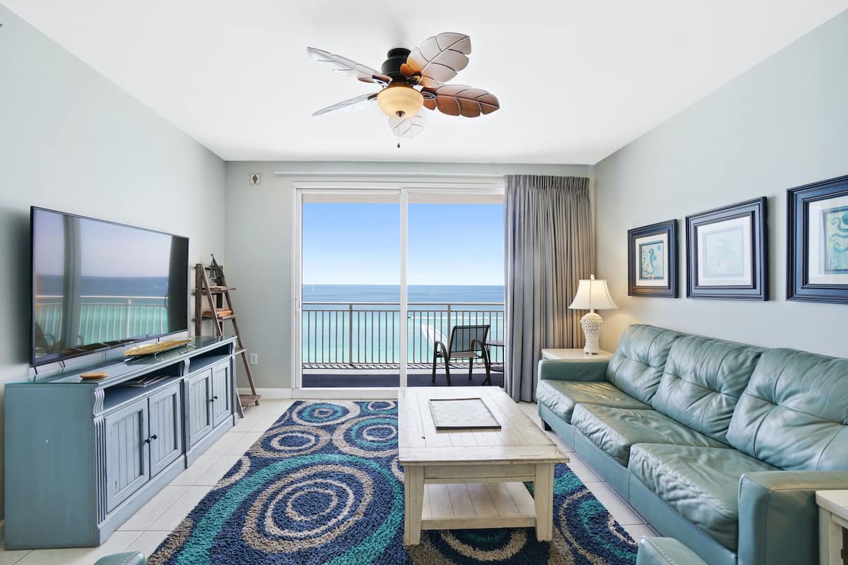 A living room with a large flat-screen TV, a green leather sofa, a wooden coffee table, and a ceiling fan.