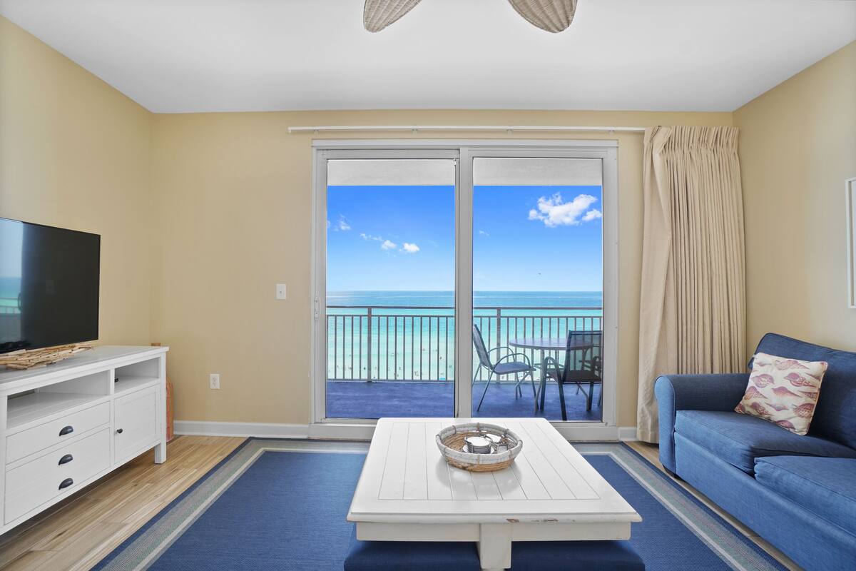 A living room with a television, a coffee table, and a sofa. There is a balcony with a view of the ocean.