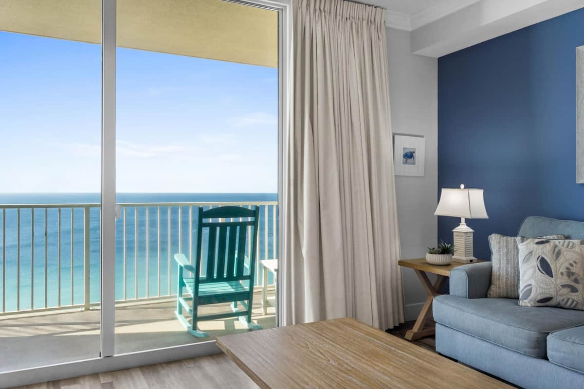 Un balcon avec vue sur l’océan, une chaise verte, une table en bois, un canapé bleu et une lampe sur une table d’appoint.