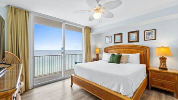 A hotel room with a large bed, a ceiling fan, a balcony with curtains, and a view of the ocean.