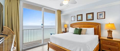 A hotel room with a large bed, a ceiling fan, a balcony with curtains, and a view of the ocean.