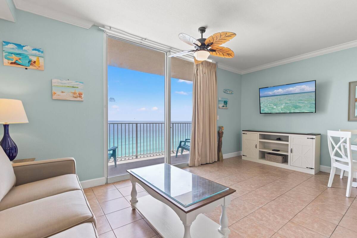 Une pièce comprenant une table avec un plateau en verre, un canapé, un téléviseur, un ventilateur de plafond, un balcon avec vue et un meuble de rangement.