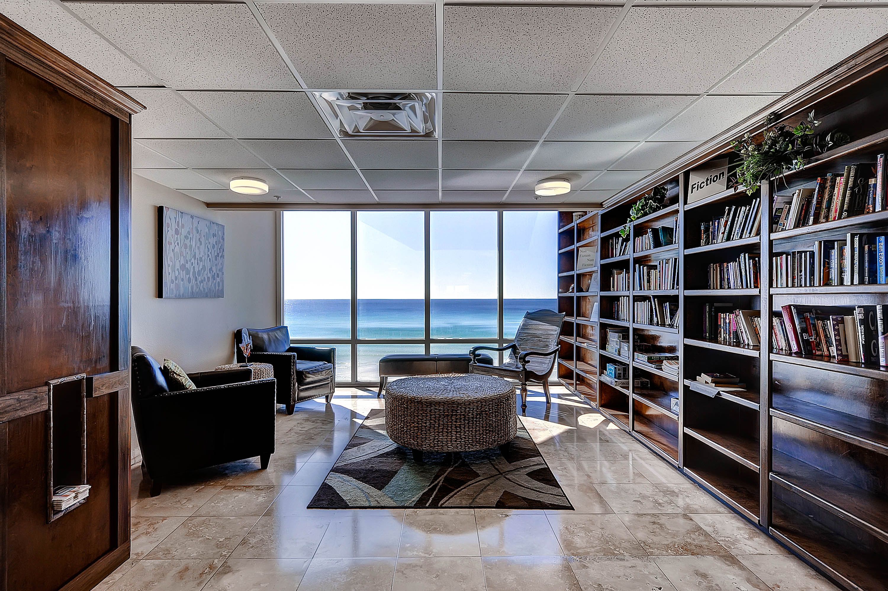 Eine Bibliothek mit großem Fenster zum Meer, ausgestattet mit Bücherregalen, einem runden Hocker und einem dunkelgrauen Sofa.