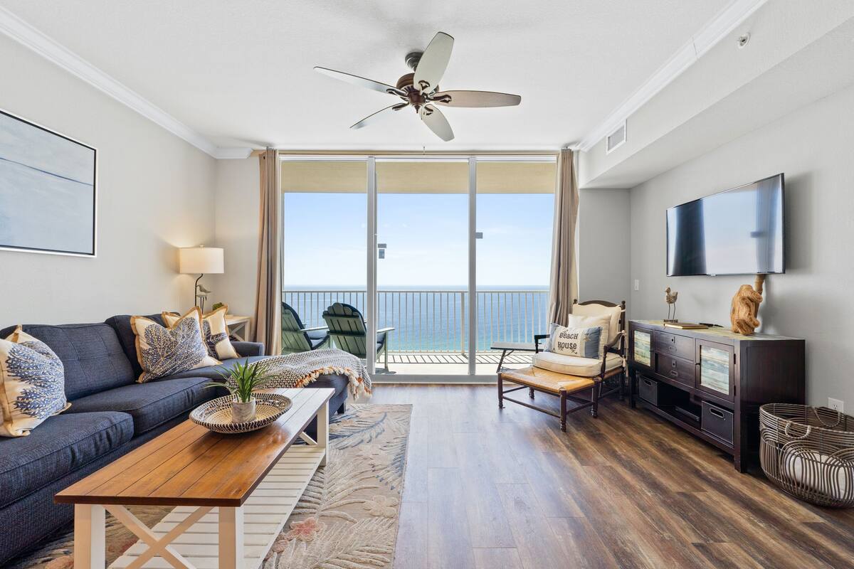 Un salon moderne avec un canapé, une table basse et un téléviseur. On peut voir un ventilateur de plafond, et une porte-fenêtre coulissante donne accès à un balcon avec vue.