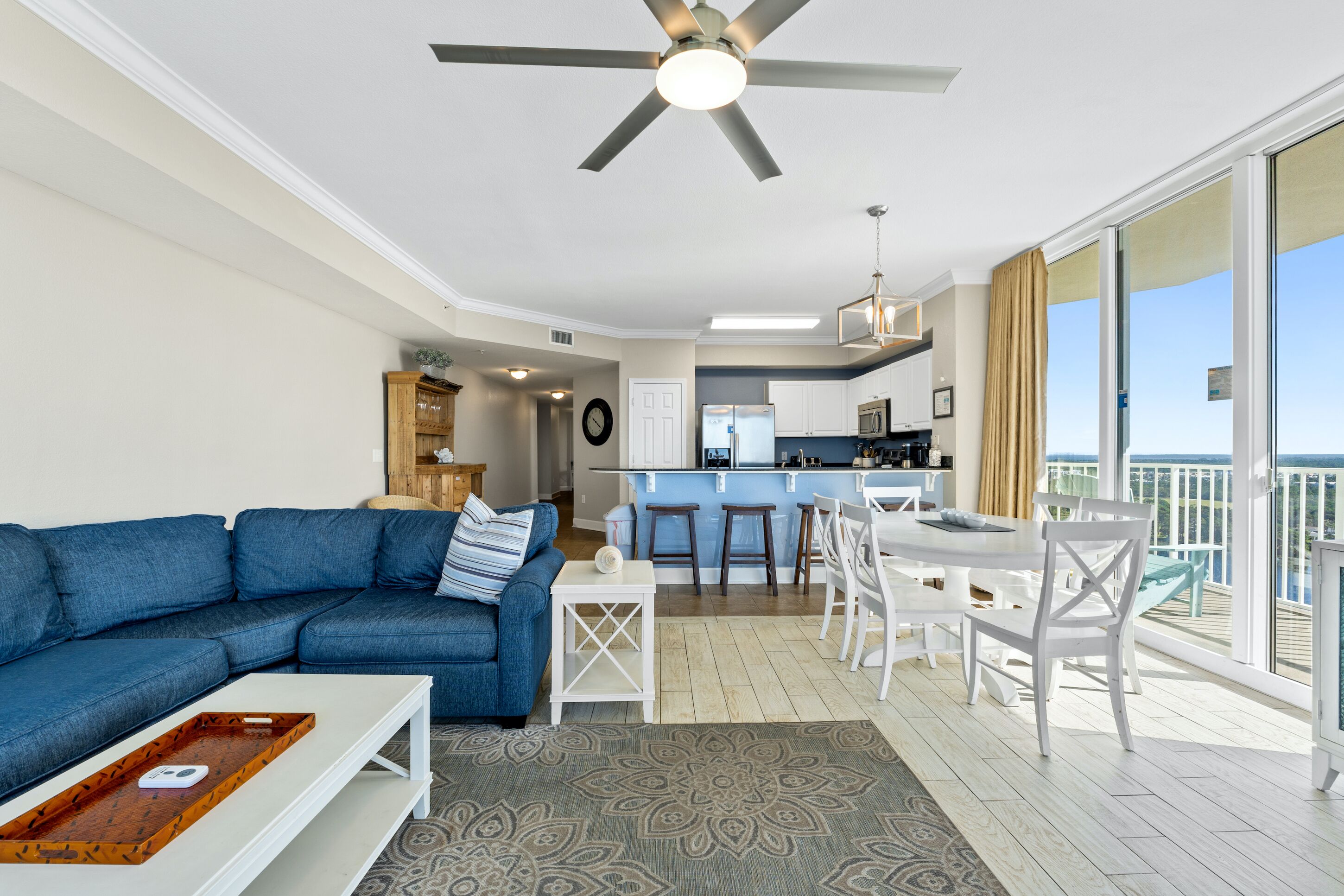 Un salon moderne avec un canapé bleu, une table basse blanche et un ventilateur de plafond. Il y a un coin cuisine avec un bar et un ensemble de salle à manger. La pièce est dotée de grandes fenêtres et d’un balcon avec vue.