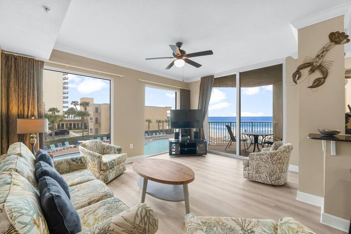 Un salon spacieux avec un grand canapé, une table basse et un téléviseur à écran plat. La pièce est dotée d’un ventilateur de plafond, de grandes fenêtres donnant sur la plage et d’un balcon avec piscine.