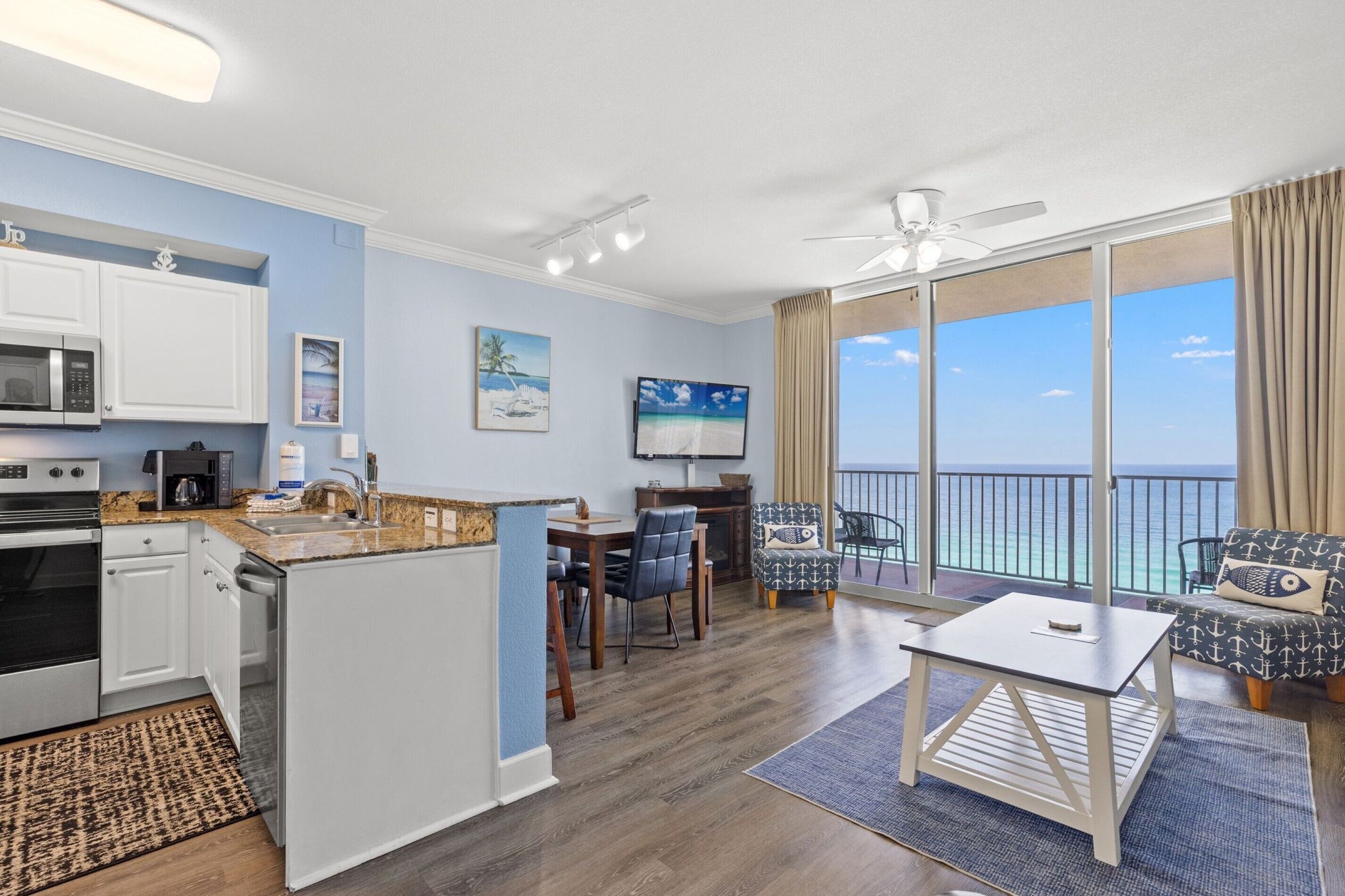 A modern kitchen with white cabinets, a microwave, and a stove. There is a dining area with a table and chairs, and a balcony with a view of the ocean.