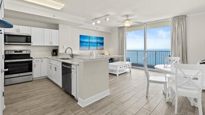 Une cuisine moderne avec des meubles blancs, un four à micro-ondes, un four traditionnel et un lave-vaisselle. On y trouve également un coin repas avec des chaises et une table blanches. Une grande fenêtre offre une vue sur l’océan.