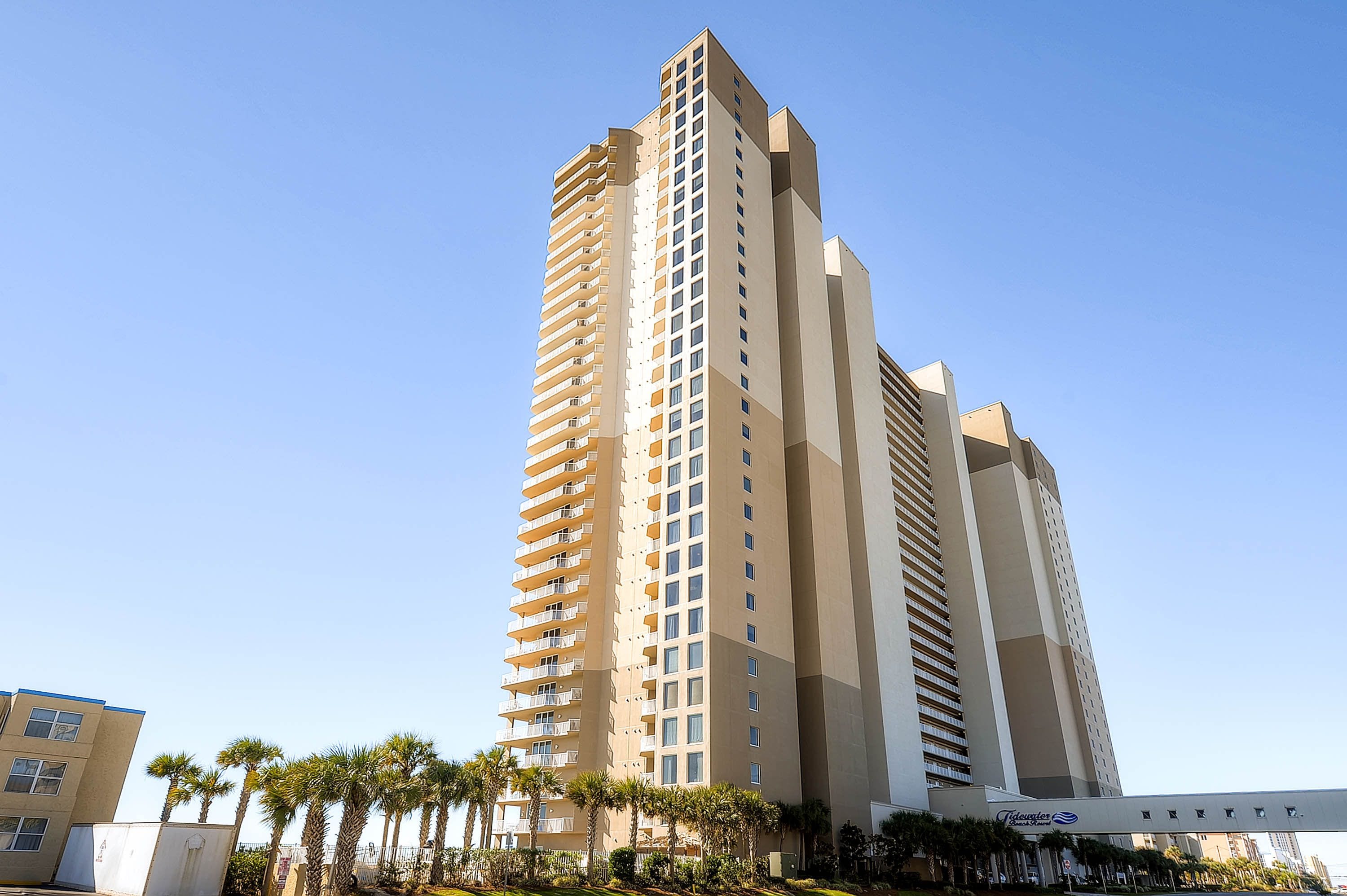 A tall, modern hotel with multiple floors and balconies, surrounded by palm trees.
