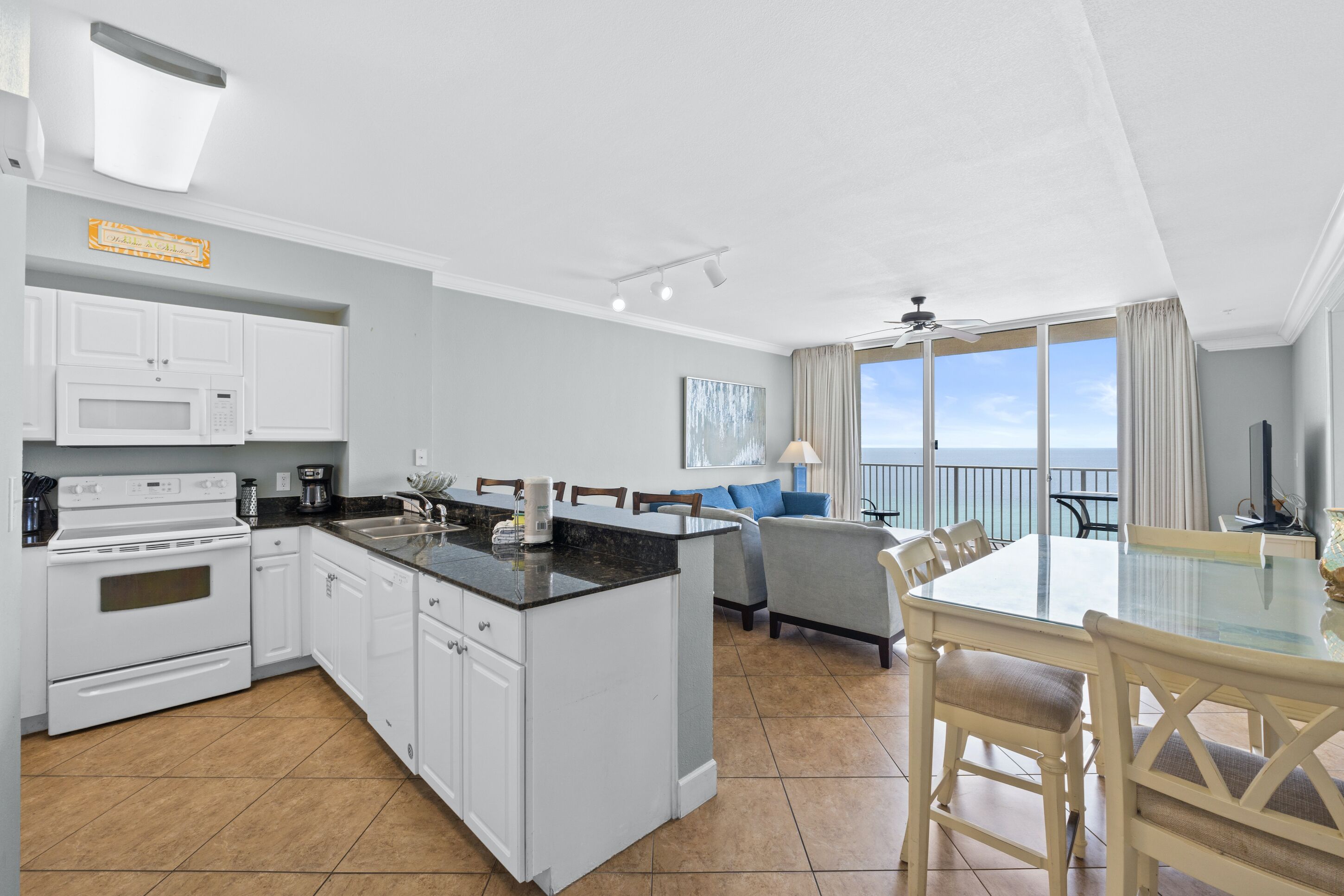 Een moderne keuken met witte kasten, een magnetron en een fornuis. Er is een eethoek met een glazen tafel en stoelen, en een groot raam met uitzicht op de oceaan.