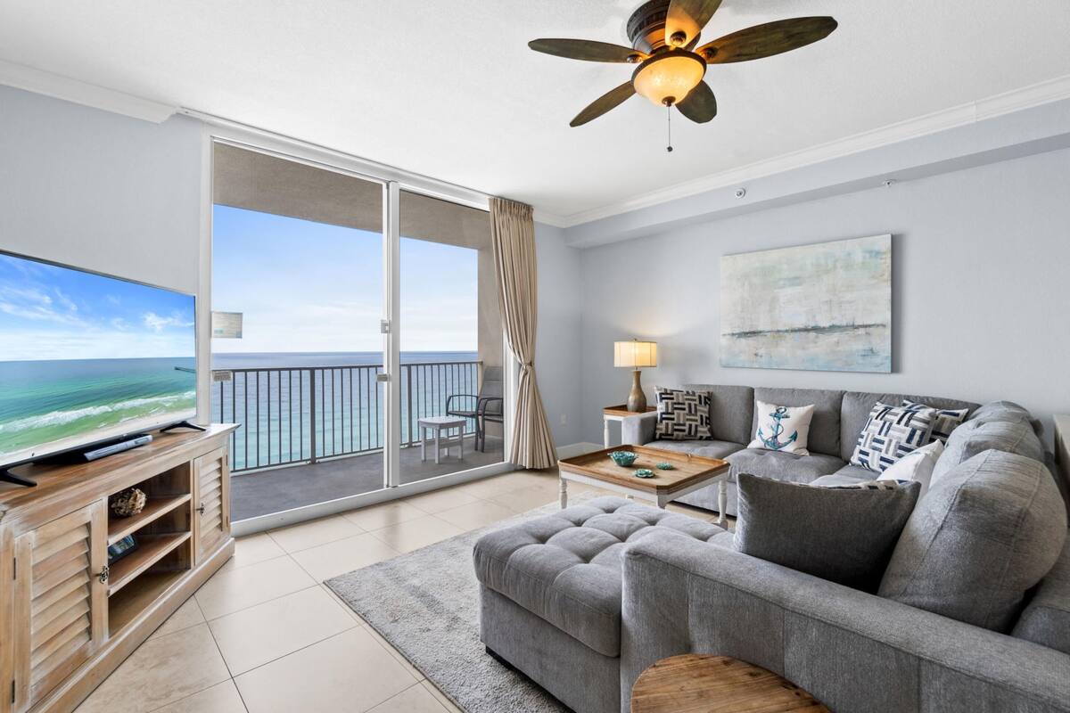 Un salon moderne avec un canapé gris, un téléviseur à écran plat, un ventilateur de plafond et un balcon offrant une vue sur l’océan.