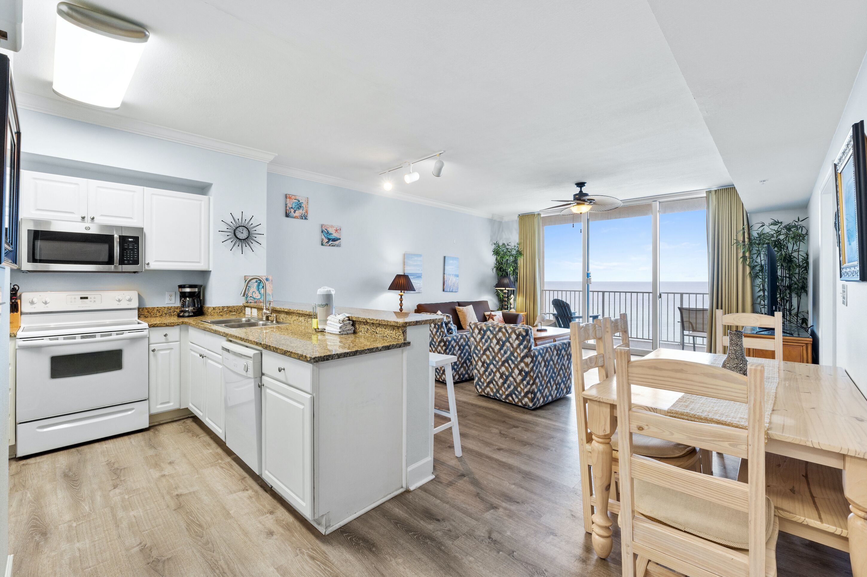Una cocina moderna con armarios blancos, microondas y estufa. Hay una zona de comedor con una mesa y sillas de madera. La habitación tiene un ventanal con vistas al océano.