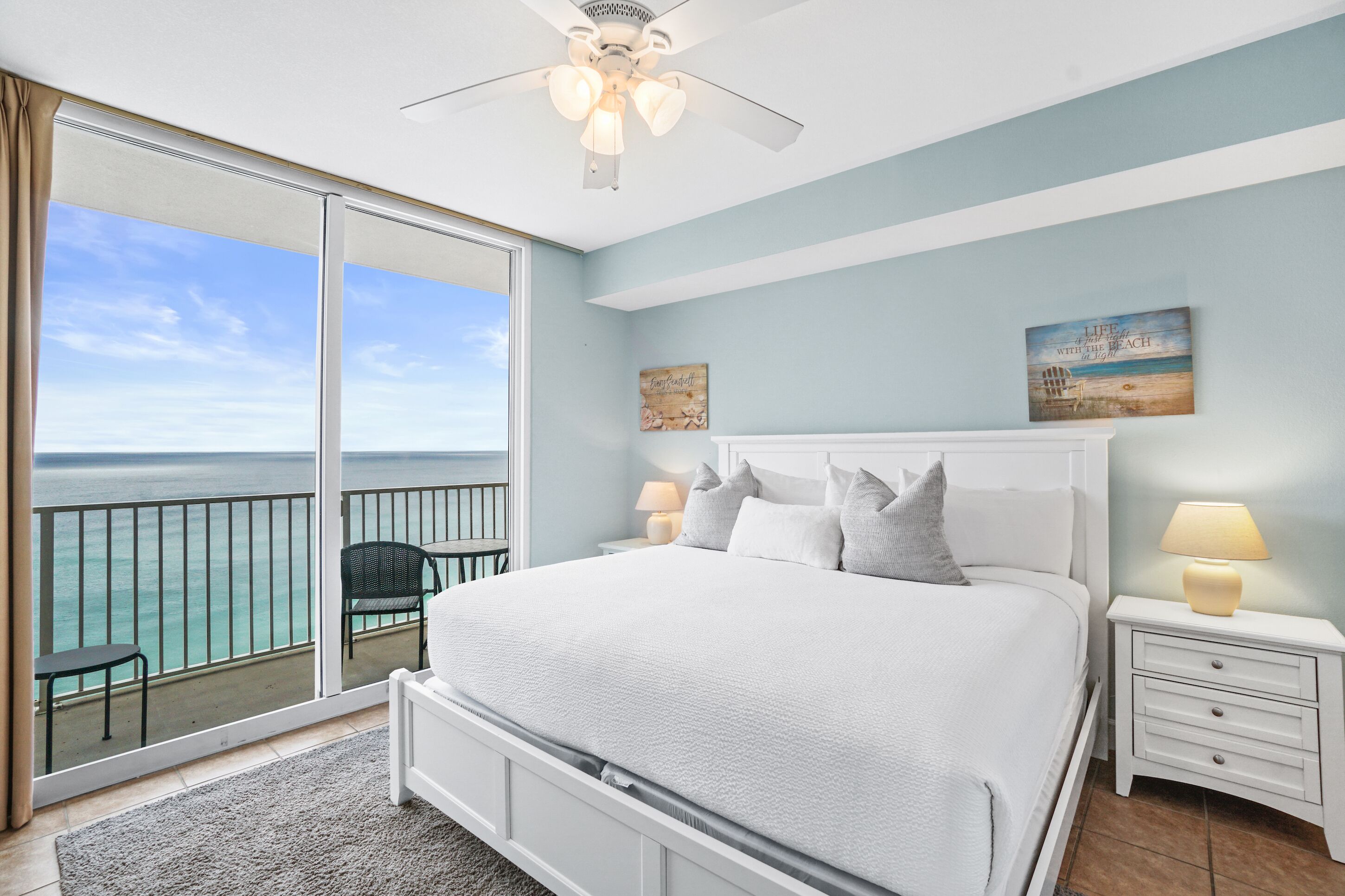A bedroom with a large bed, a ceiling fan, a nightstand with a lamp, and a view of the ocean through a sliding glass door.
