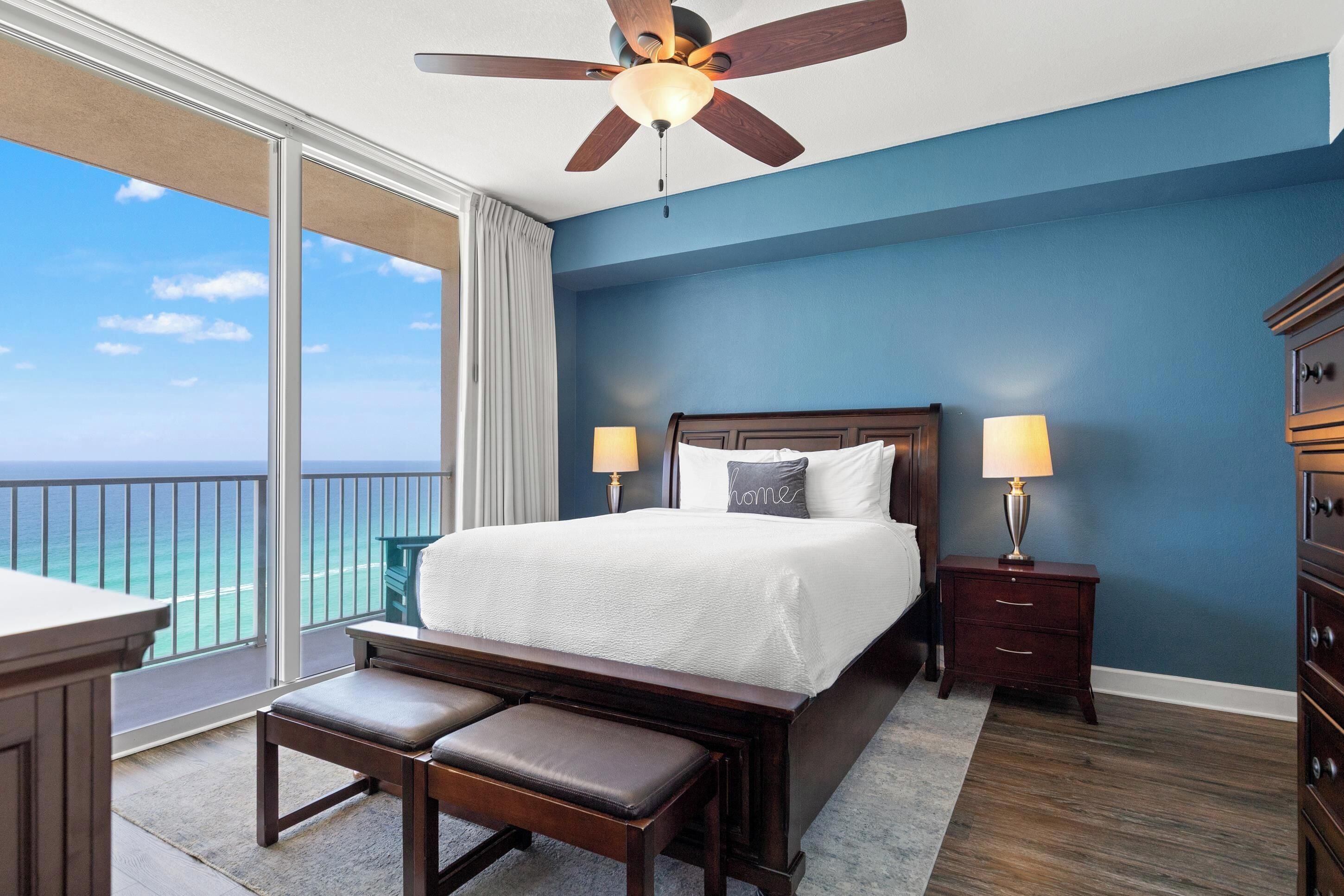 A hotel room with a large bed, two bedside tables with lamps, a bench, and a balcony with a view of the ocean.