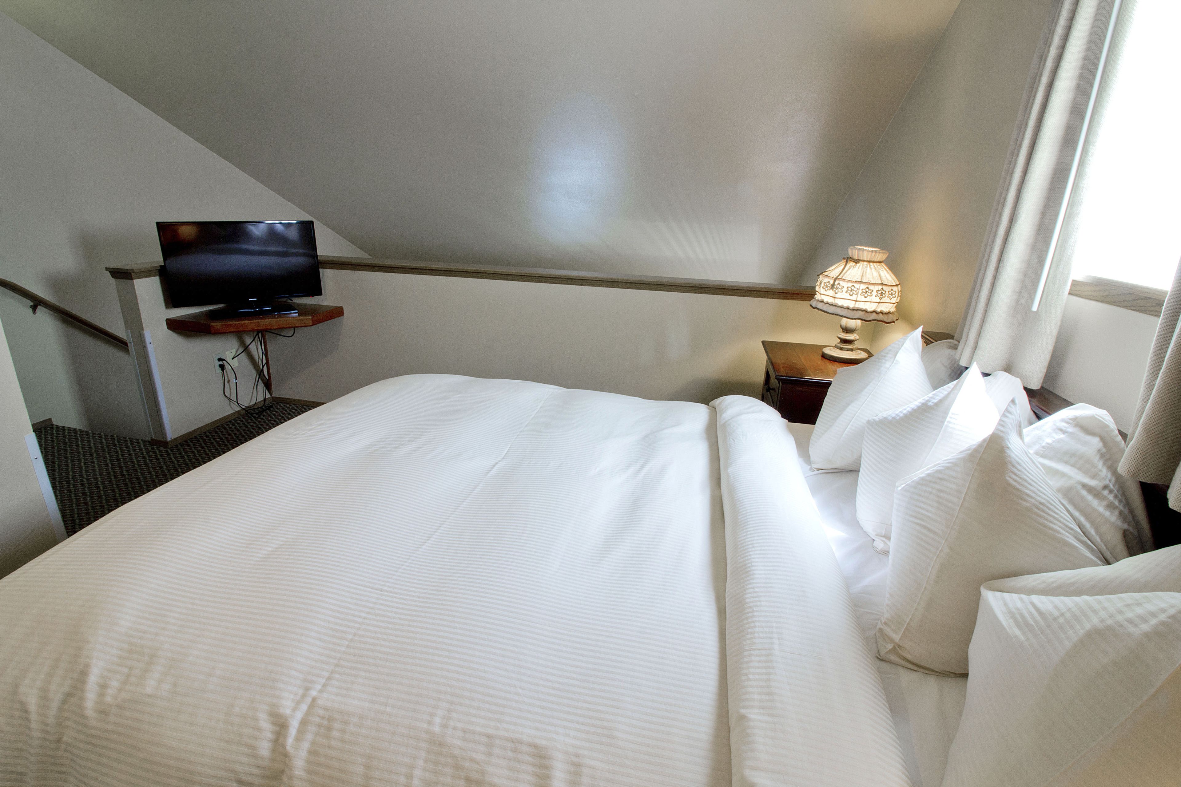 A hotel room with a bed, a television on a stand, a nightstand with a lamp, and a window with curtains.