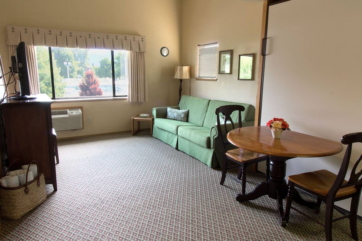 A room with a green sofa, a wooden dining table with chairs, a lamp, and a window with curtains.