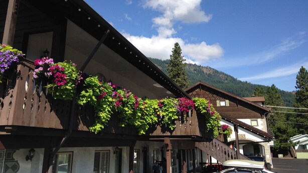 Un chalet traditionnel avec des balcons en bois ornés de fleurs, un paysage montagneux en toile de fond et un ciel bleu azur.