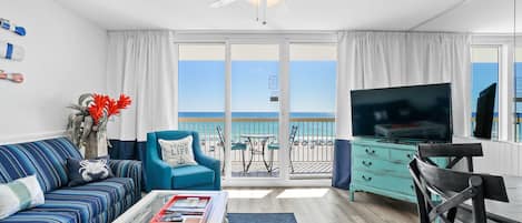 A living room with a sofa, armchair, TV, and a balcony with a view of the beach.