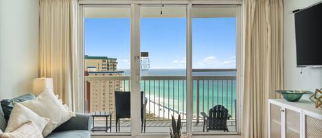 A living room with a sofa, a coffee table, and a view of the beach and ocean through a sliding glass door.
