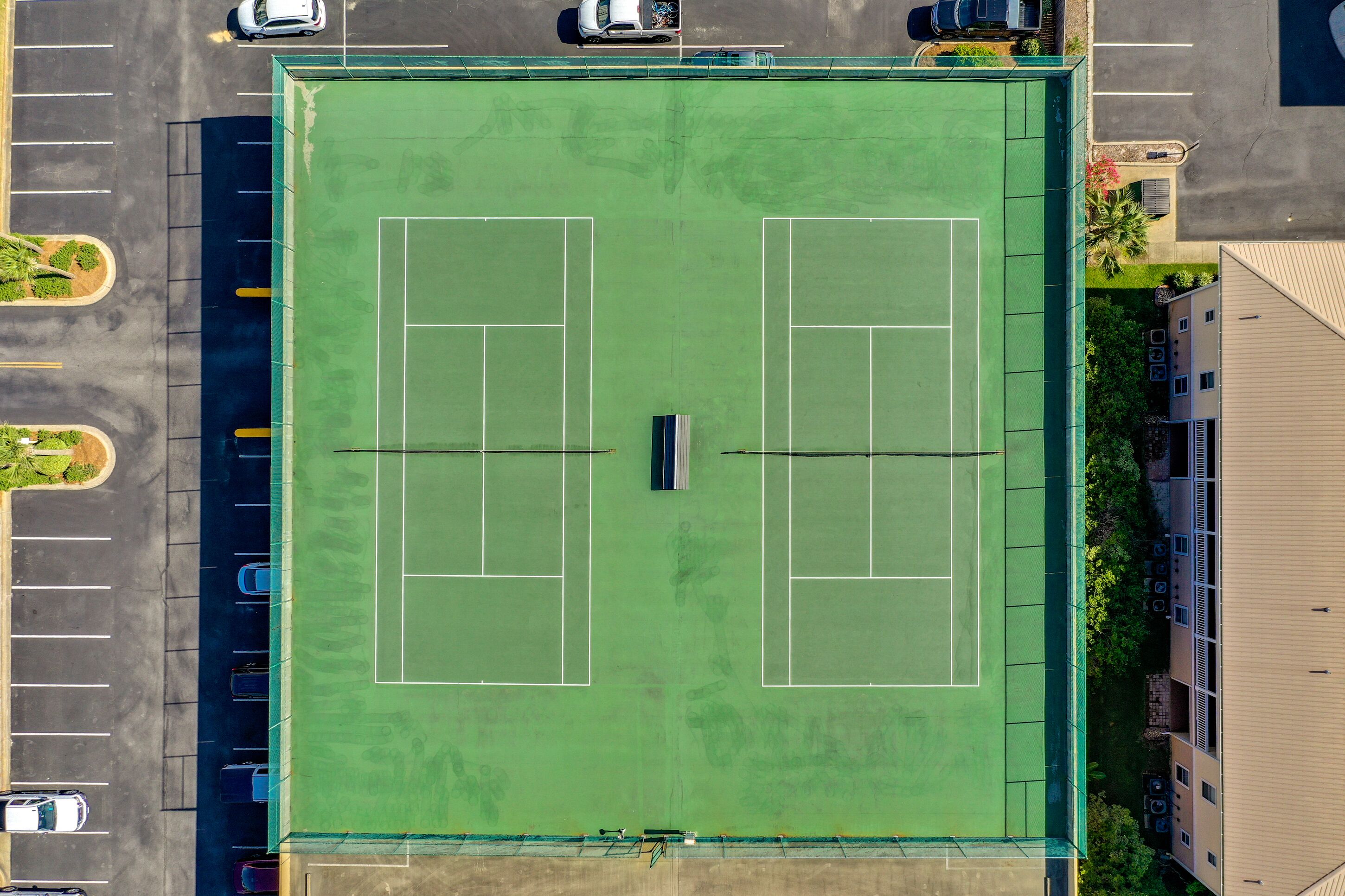 Vista aérea de una cancha de tenis rodeada por un estacionamiento y edificios.