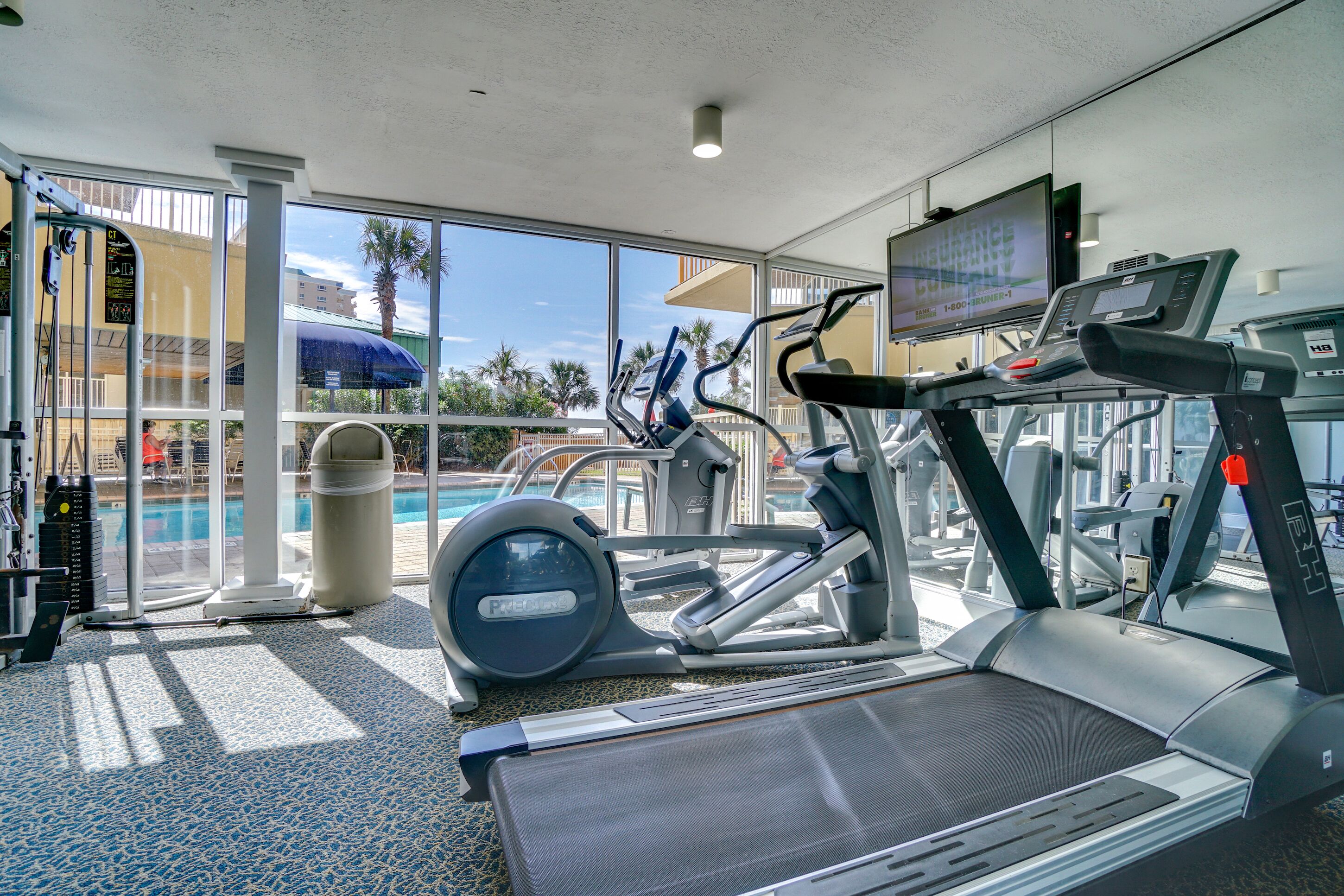 A gym with treadmills, elliptical machines, and a pool area visible through large windows.