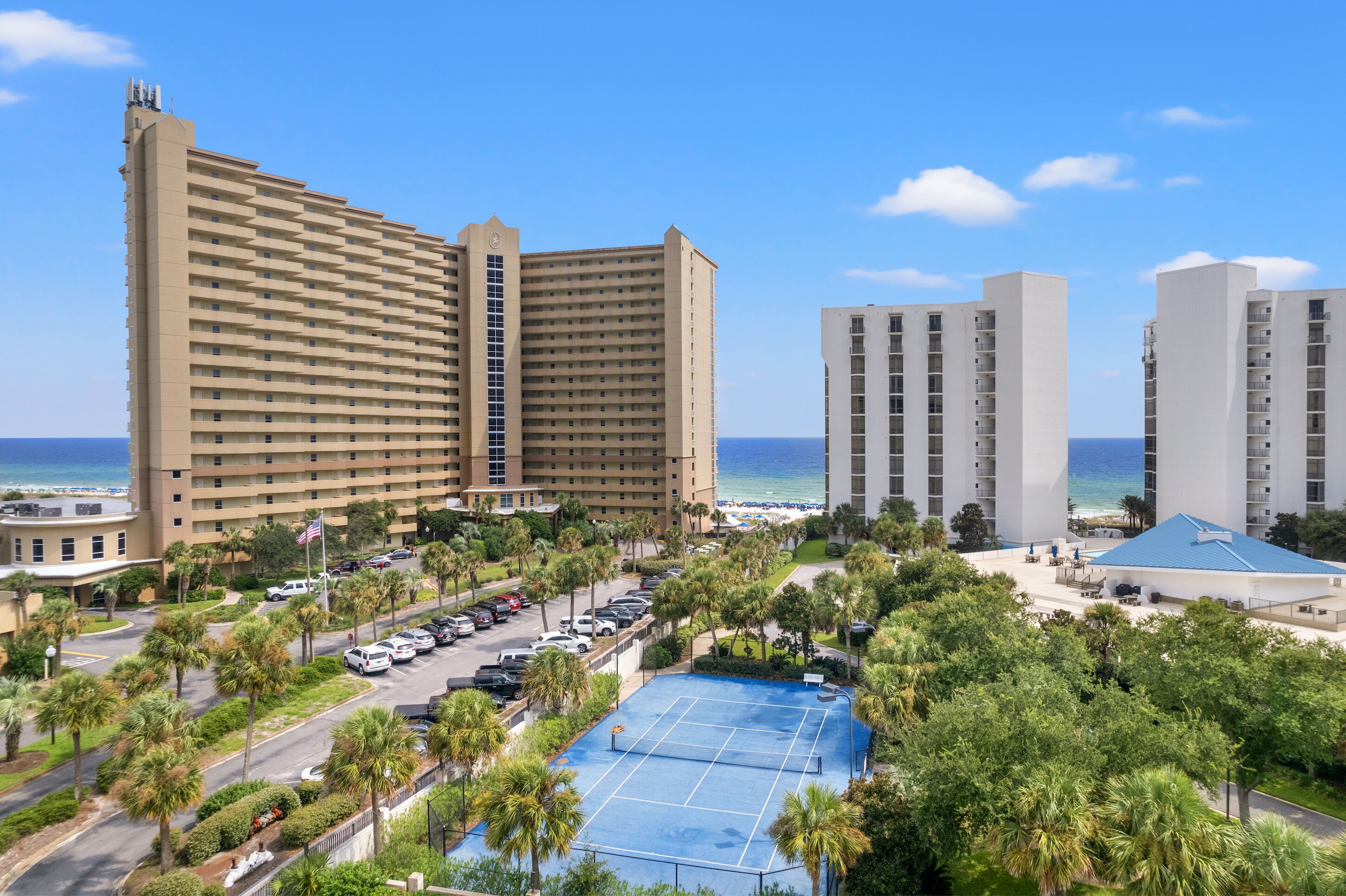A hotel with multiple high-rise buildings, a parking lot, and a tennis court surrounded by palm trees.