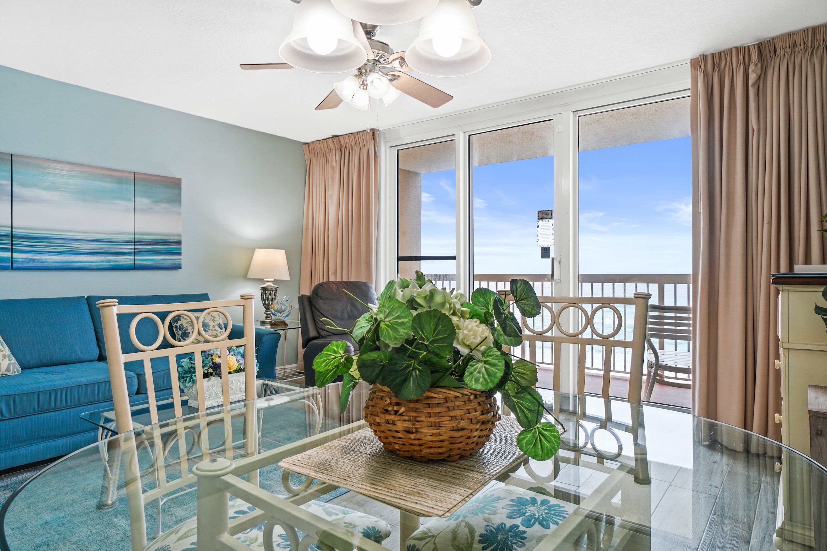A living room with a blue sofa, a glass coffee table with a wicker centerpiece, a ceiling fan, and a view of the outdoors through a sliding glass door.