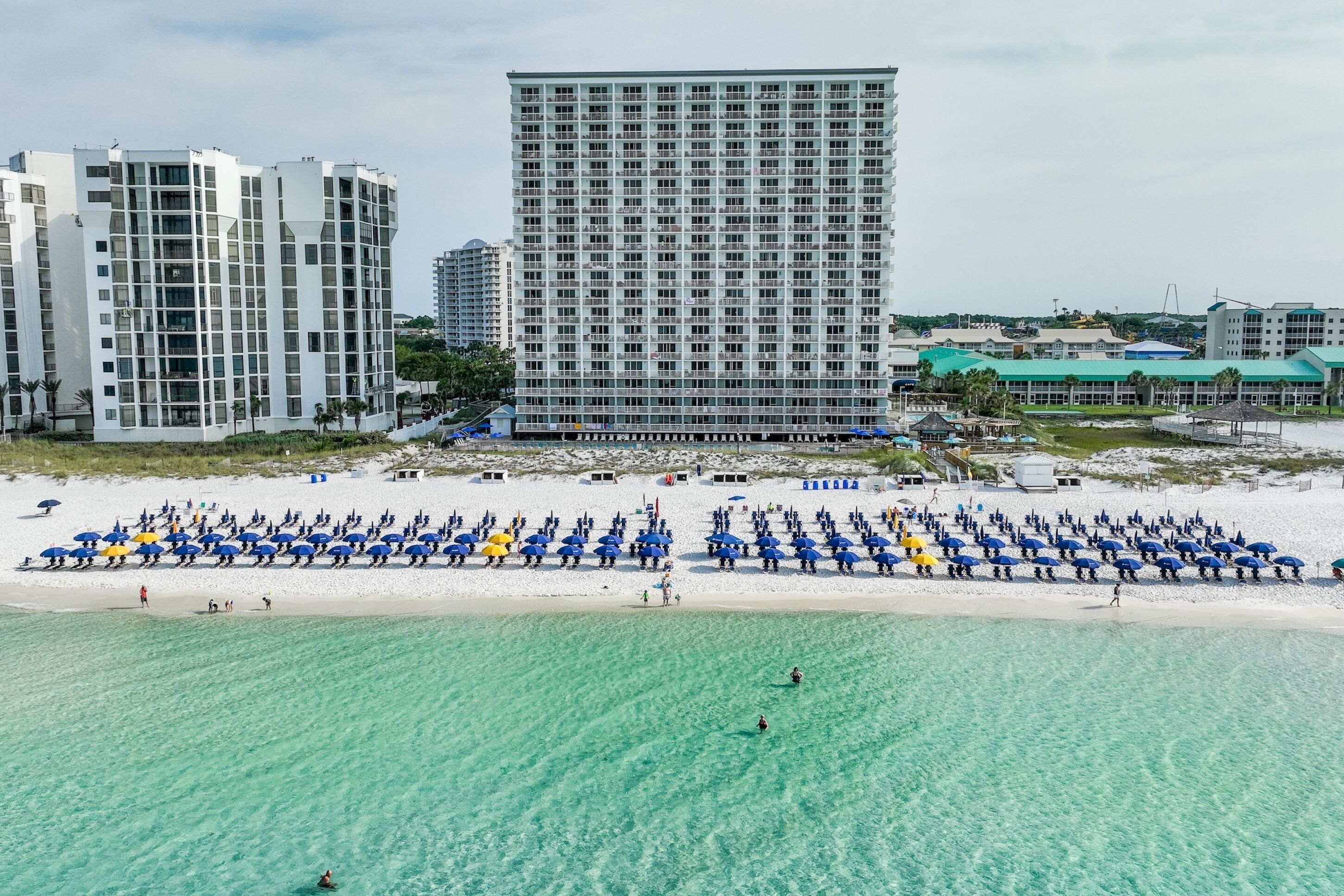 A beachfront hotel with a large building, a sandy beach with umbrellas, and clear turquoise water.