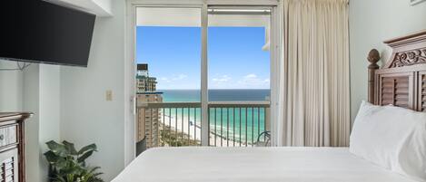 Habitación de hotel con una cama grande, ventilador de techo, televisor, planta en maceta y balcón con vista a la playa y al océano.