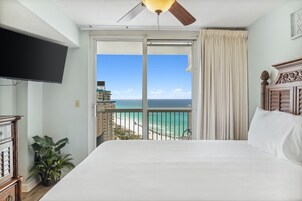 Habitación de hotel con una cama grande, ventilador de techo, televisor, planta en maceta y balcón con vista a la playa y al océano.
