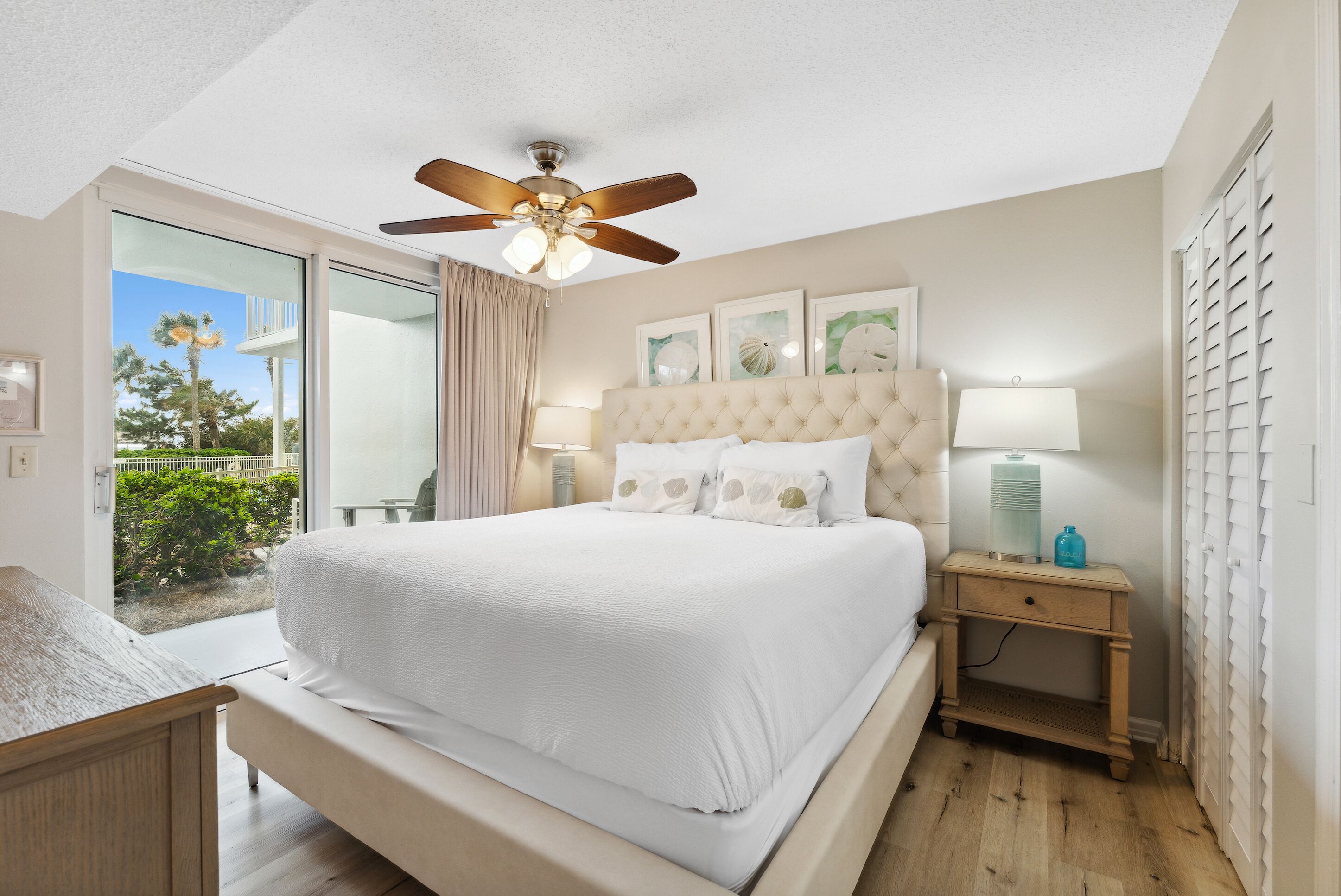 A bedroom with a large bed, a ceiling fan, a nightstand, and a view of the outdoors.