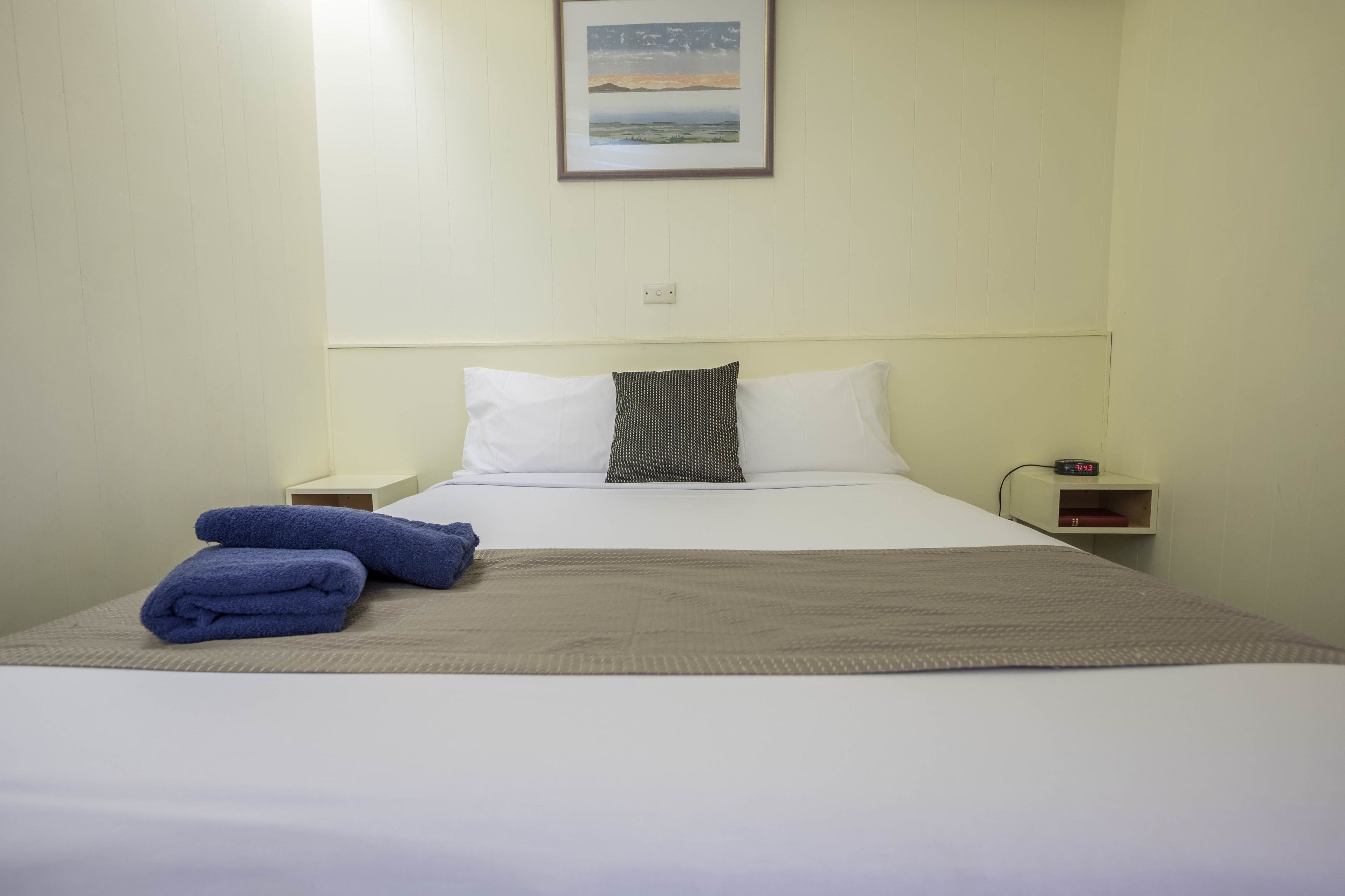 Habitación de hotel con cama, almohadas, manta y mesita de noche con un reloj.